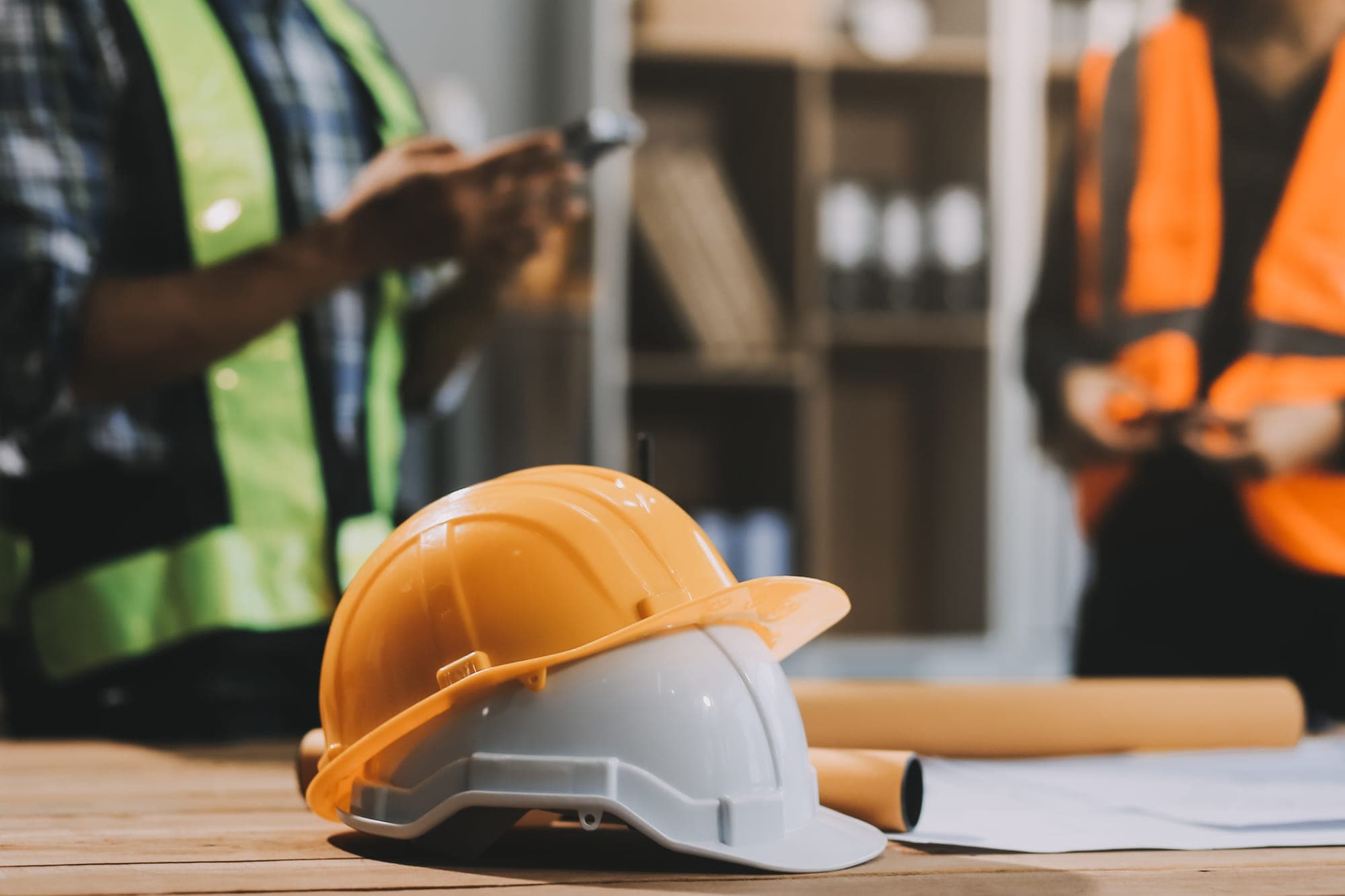 Deux casques de chantier posés sur des plans, une équipe en gilets en arrière-plan